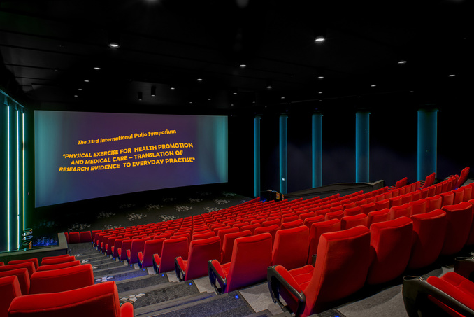The picture of the auditorium of Finnkino Scala with the text reflected on its screen: "International 23rd Puijo Symposium - "Physical Exercise for Health Promotion and Medical Care - Translation of Research Evidence to Everyday Practice".